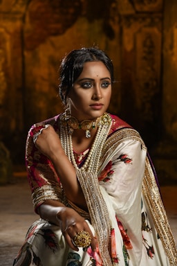 A woman is dressed in traditional attire, featuring a richly embroidered sari with floral patterns and golden accents. She is adorned with elaborate jewelry, including a choker necklace with pearls and a large ring. The background is dimly lit, adding a dramatic effect, emphasizing her poised and elegant demeanor.