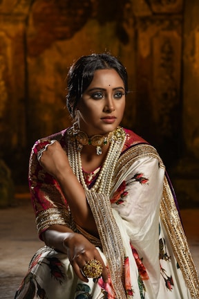 A woman is dressed in traditional attire, featuring a richly embroidered sari with floral patterns and golden accents. She is adorned with elaborate jewelry, including a choker necklace with pearls and a large ring. The background is dimly lit, adding a dramatic effect, emphasizing her poised and elegant demeanor.
