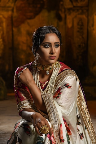 A woman is dressed in traditional attire, featuring a richly embroidered sari with floral patterns and golden accents. She is adorned with elaborate jewelry, including a choker necklace with pearls and a large ring. The background is dimly lit, adding a dramatic effect, emphasizing her poised and elegant demeanor.
