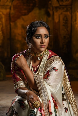 A woman is dressed in traditional attire, featuring a richly embroidered sari with floral patterns and golden accents. She is adorned with elaborate jewelry, including a choker necklace with pearls and a large ring. The background is dimly lit, adding a dramatic effect, emphasizing her poised and elegant demeanor.