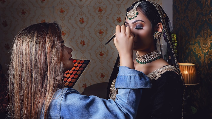 A makeup artist applies eye makeup to a woman dressed in traditional attire with intricate jewelry. The setting includes a warmly lit room with patterned wallpaper and a floor lamp, adding to the aesthetic ambiance.