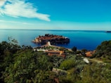 An island connected to the mainland by a narrow causeway is surrounded by calm blue waters. The island features historic stone buildings and lush greenery, creating a picturesque scene. The mainland foreground includes dense vegetation and a few buildings with terracotta roofs.