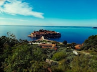 An island connected to the mainland by a narrow causeway is surrounded by calm blue waters. The island features historic stone buildings and lush greenery, creating a picturesque scene. The mainland foreground includes dense vegetation and a few buildings with terracotta roofs.