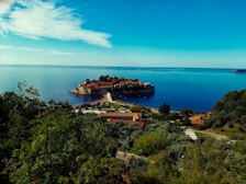 An island connected to the mainland by a narrow causeway is surrounded by calm blue waters. The island features historic stone buildings and lush greenery, creating a picturesque scene. The mainland foreground includes dense vegetation and a few buildings with terracotta roofs.