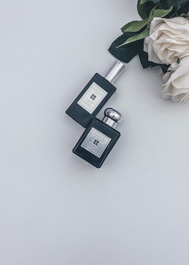 Two black perfume bottles are positioned on a white surface, accompanied by a cluster of cream-colored roses with dark green leaves.