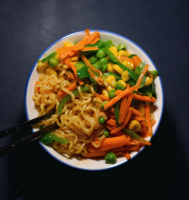 A bowl filled with instant noodles topped with a colorful array of vegetables, including sliced carrots, green bell peppers, peas, and corn. The dish is neatly arranged, with chopsticks resting on the side, creating a visually appealing and appetizing meal.