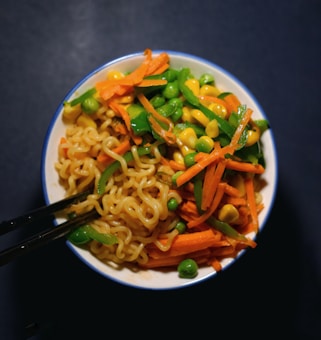A bowl filled with instant noodles topped with a colorful array of vegetables, including sliced carrots, green bell peppers, peas, and corn. The dish is neatly arranged, with chopsticks resting on the side, creating a visually appealing and appetizing meal.