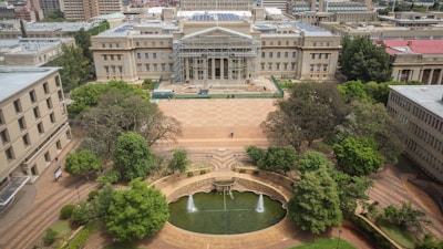 An aerial view of a large, historic academic building with a grand entrance featuring columns. The building is under partial renovation with scaffolding visible. In the foreground, a landscaped area with trees, bushes, and a circular pond featuring fountains. The surroundings include other architectural structures, suggesting a university campus.
