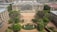 An aerial view of a large, historic academic building with a grand entrance featuring columns. The building is under partial renovation with scaffolding visible. In the foreground, a landscaped area with trees, bushes, and a circular pond featuring fountains. The surroundings include other architectural structures, suggesting a university campus.
