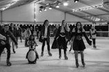A group of people ice skating on an indoor rink, with one child using a penguin skating aid. The rink is well-lit with hanging lights and a disco ball. Some skaters are dressed in costumes, suggesting a themed event.