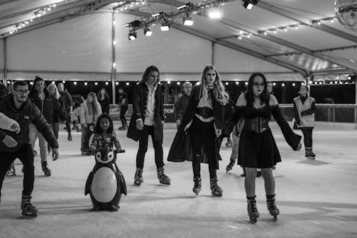 A group of people ice skating on an indoor rink, with one child using a penguin skating aid. The rink is well-lit with hanging lights and a disco ball. Some skaters are dressed in costumes, suggesting a themed event.
