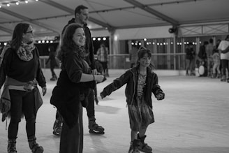 Several people are enjoying ice skating indoors under string lights in a covered rink. A young girl with face paint is skating happily with adults nearby, all wearing ice skates and casual winter attire, with expressions of joy and amusement.