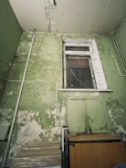 A room with deteriorating green walls showing signs of peeling paint and cracked plaster. There is a wooden cupboard or cabinet against the wall with a transparent container on top. Above it is a sealed window with a frame that also shows signs of wear and tear. Various pipes run along the walls and ceiling.
