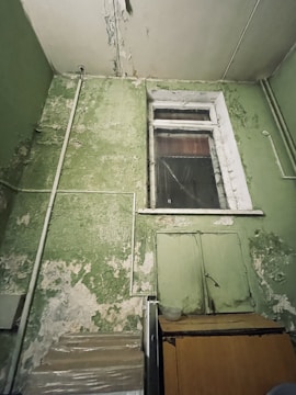 A room with deteriorating green walls showing signs of peeling paint and cracked plaster. There is a wooden cupboard or cabinet against the wall with a transparent container on top. Above it is a sealed window with a frame that also shows signs of wear and tear. Various pipes run along the walls and ceiling.