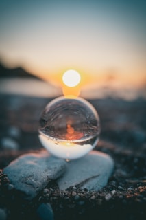 A crystal ball positioned on a rocky surface reflects a serene sunset over a body of water. The sunlight creates a warm glow and bokeh effect in the background, blurring into a gradient sky from blue to orange.