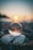 A crystal ball positioned on a rocky surface reflects a serene sunset over a body of water. The sunlight creates a warm glow and bokeh effect in the background, blurring into a gradient sky from blue to orange.