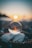 A crystal ball positioned on a rocky surface reflects a serene sunset over a body of water. The sunlight creates a warm glow and bokeh effect in the background, blurring into a gradient sky from blue to orange.