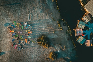 Aerial view of a beach setting featuring colorful sunbeds arranged in a somewhat circular pattern with people lounging. Long shadows are cast on the sand, indicating late afternoon or early morning sunlight. To the right, there are structures with colorful roofs, possibly beach huts or stalls, surrounded by dark sand or soil.