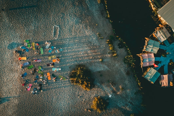 Aerial view of a beach setting featuring colorful sunbeds arranged in a somewhat circular pattern with people lounging. Long shadows are cast on the sand, indicating late afternoon or early morning sunlight. To the right, there are structures with colorful roofs, possibly beach huts or stalls, surrounded by dark sand or soil.