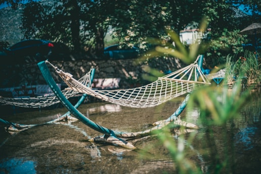 A netted hammock is suspended over a shallow body of water, surrounded by lush greenery. The background consists of trees and a stone wall with shadows cast by the sunlight. The scene conveys a sense of tranquility and relaxation, perfect for a peaceful retreat in nature.