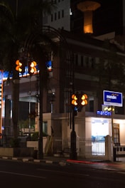 A nighttime urban scene featuring a well-lit street corner with a bank ATM. The area includes traffic lights and a palm tree, with a building in the background. There is a distinct contrast between the illuminated signs and the dark street.