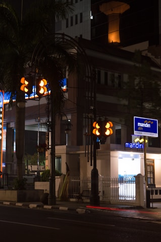 A nighttime urban scene featuring a well-lit street corner with a bank ATM. The area includes traffic lights and a palm tree, with a building in the background. There is a distinct contrast between the illuminated signs and the dark street.