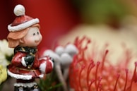 A cute figurine of a child dressed in a red and white winter outfit is smiling. The child holds a small, red-and-white object, and is surrounded by vibrant, detailed flowers. The background includes blurred green and red elements, adding to the festive theme.
