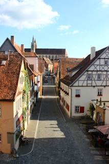 A picturesque cobblestone street in a European village flanked by charming, rustic buildings with timber framing and red-tiled roofs. The narrow street, adorned with flowering plants, leads to a magnificent church in the background. A few tables with people seated and a bicycle leaning against a wall add to the quaint atmosphere.