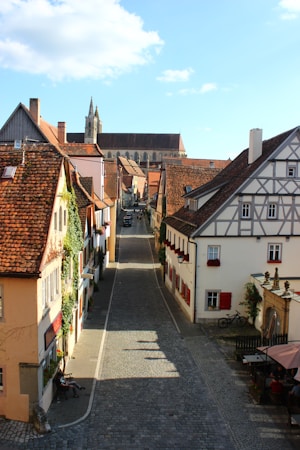 A picturesque cobblestone street in a European village flanked by charming, rustic buildings with timber framing and red-tiled roofs. The narrow street, adorned with flowering plants, leads to a magnificent church in the background. A few tables with people seated and a bicycle leaning against a wall add to the quaint atmosphere.