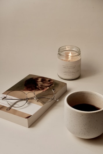 A cozy, minimalistic scene featuring a closed book with a pair of round glasses resting on it, placed beside a lit candle and a speckled ceramic mug filled with a dark beverage. The items are arranged on a smooth light surface.