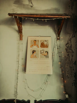 A wall-mounted calendar with illustrations hangs under a wooden shelf. The calendar is surrounded by a string of white fairy lights draped in a U-shape. The wall appears textured and slightly worn, contributing to a rustic atmosphere.