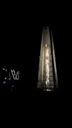 A tall, pyramid-shaped outdoor patio heater is illuminated against a dark background. The heater emits a bright, warm light from the fire inside its glass enclosure, creating a striking contrast with the surrounding darkness.