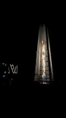 A tall, pyramid-shaped outdoor patio heater is illuminated against a dark background. The heater emits a bright, warm light from the fire inside its glass enclosure, creating a striking contrast with the surrounding darkness.