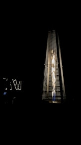 A tall, pyramid-shaped outdoor patio heater is illuminated against a dark background. The heater emits a bright, warm light from the fire inside its glass enclosure, creating a striking contrast with the surrounding darkness.