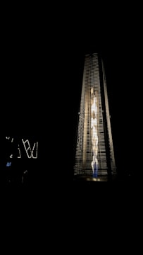 A tall, pyramid-shaped outdoor patio heater is illuminated against a dark background. The heater emits a bright, warm light from the fire inside its glass enclosure, creating a striking contrast with the surrounding darkness.