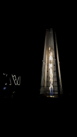 A tall, pyramid-shaped outdoor patio heater is illuminated against a dark background. The heater emits a bright, warm light from the fire inside its glass enclosure, creating a striking contrast with the surrounding darkness.