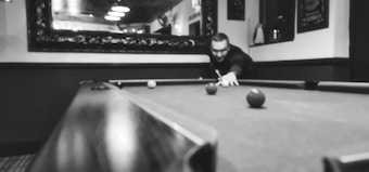 A person is playing pool on a billiard table, focusing on hitting the cue ball. The scene is indoors, possibly in a bar or game room, with a large ornate mirror on the wall reflecting ceiling lights.