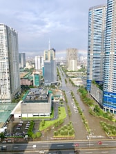 A flooded urban street surrounded by tall skyscrapers and modern buildings, with water covering several lanes. The scene includes a rainbow visible in the distance, indicating a recent rainfall. Vehicles and people navigate the partially submerged cityscape, and trees line the avenues.