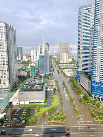 A flooded urban street surrounded by tall skyscrapers and modern buildings, with water covering several lanes. The scene includes a rainbow visible in the distance, indicating a recent rainfall. Vehicles and people navigate the partially submerged cityscape, and trees line the avenues.