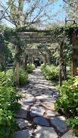 A serene garden pathway adorned with flat stone tiles, surrounded by lush greenery. Wooden pergolas covered in climbing plants create a tunnel-like effect, while dappled sunlight filters through the leaves. The scene conveys a sense of tranquility and natural beauty.