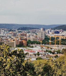 A bustling urban landscape with numerous buildings of varying heights, surrounded by lush greenery in the foreground. Construction cranes are visible, indicating development activity. Hills can be seen in the background under a partly cloudy sky.