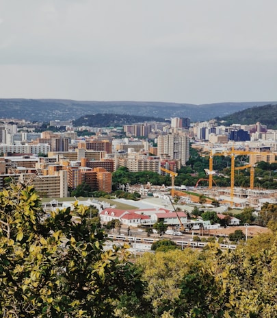 A bustling urban landscape with numerous buildings of varying heights, surrounded by lush greenery in the foreground. Construction cranes are visible, indicating development activity. Hills can be seen in the background under a partly cloudy sky.