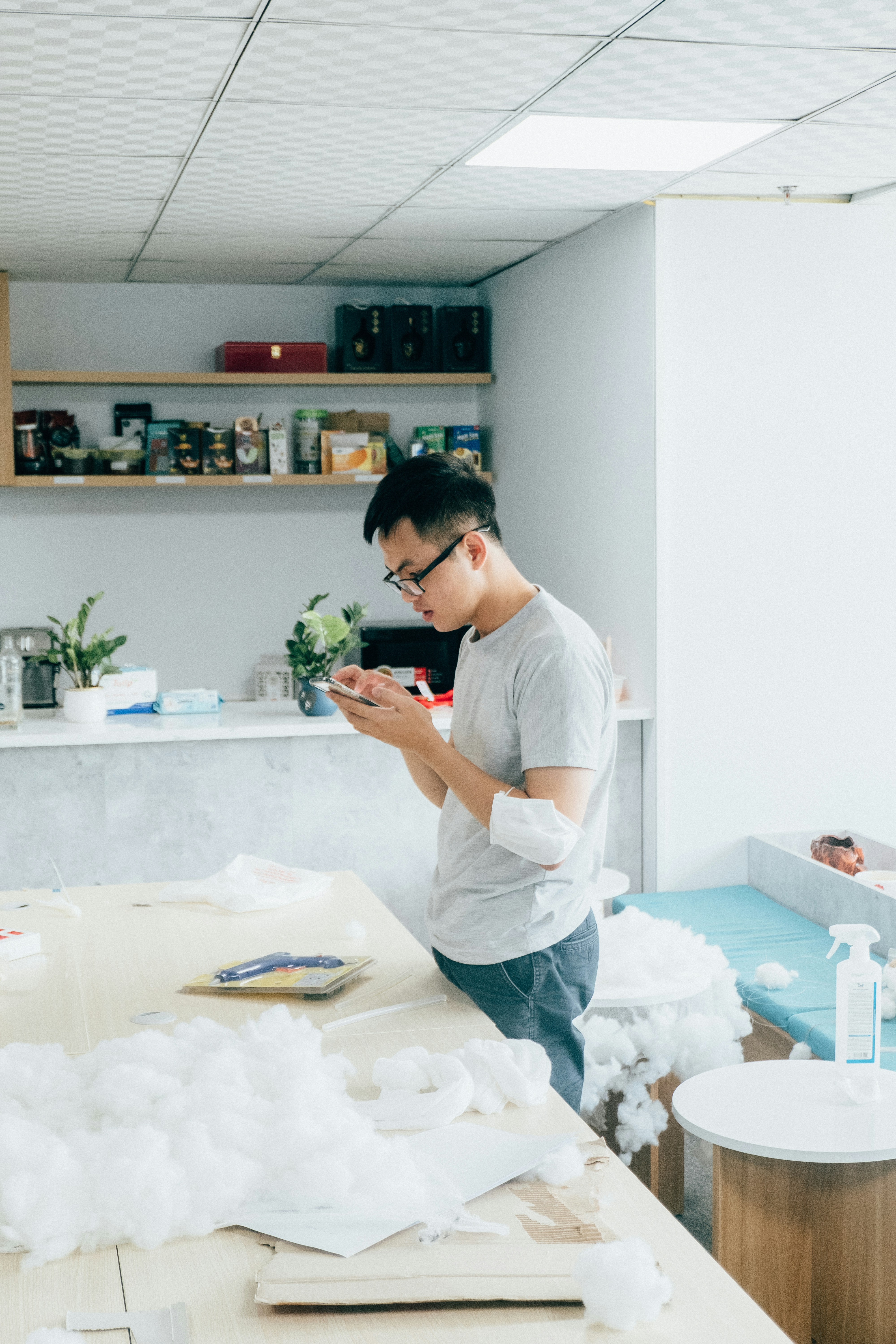 Un homme en t-shirt gris et lunettes, debout devant un comptoir, regarde l'écran de son téléphone, avec des plantes et des étagères derrière lui