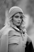 A person wearing a cozy coat and a knitted beanie looks directly into the camera. The setting is outdoors, with a blurred background suggesting a natural environment. The image is in black and white, emphasizing the textures and contrasts in the clothing and the person's features.
