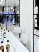 A modern indoor space with a long white table and beige chairs. Several beer bottles are placed in an organized manner on the table, alongside a small spray bottle and some utensils. Visible through a glass partition, a group of people is seen interacting in a hallway with glossy tiled flooring and decorative round lights.