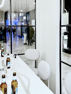 A modern indoor space with a long white table and beige chairs. Several beer bottles are placed in an organized manner on the table, alongside a small spray bottle and some utensils. Visible through a glass partition, a group of people is seen interacting in a hallway with glossy tiled flooring and decorative round lights.