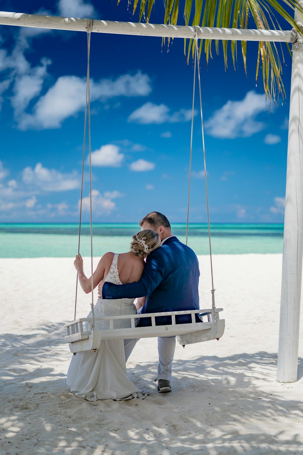 a bride and groom sitting on a swing at the beach