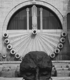 An architectural structure with a large stone or concrete head sculpture in the foreground. Above the sculpture is a circular window with intricate designs including radial lines and rosettes. The design is symmetrical with a classical style, featuring columns and ornate patterns.