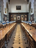 A grand dining hall with long wooden tables lined with chairs, set under a high ceiling. The walls are adorned with large portraits in ornate frames. Elegant blue-shaded table lamps are evenly spaced along the tables, and the floor is patterned with black and white tiles.