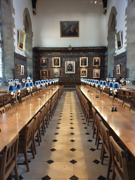 A grand dining hall with long wooden tables lined with chairs, set under a high ceiling. The walls are adorned with large portraits in ornate frames. Elegant blue-shaded table lamps are evenly spaced along the tables, and the floor is patterned with black and white tiles.